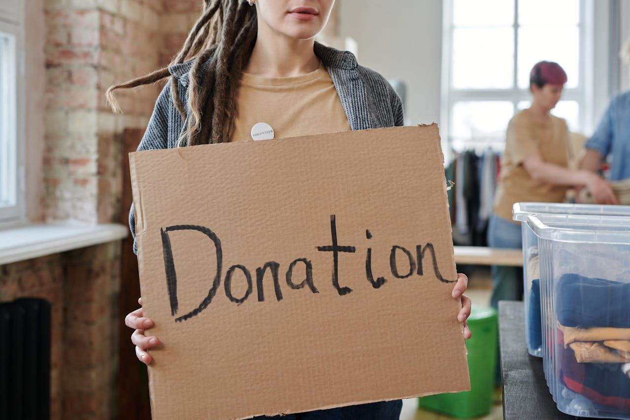 Volunteer holding a cardboard donation sign in a community center with clothing and supplies.